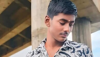 Young man with short black hair looking down, wearing a patterned shirt, under a concrete structure.