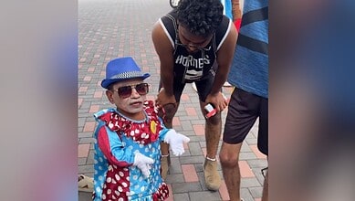 Child dressed as a clown with sunglasses and a blue hat, standing outdoors on a brick pavement.