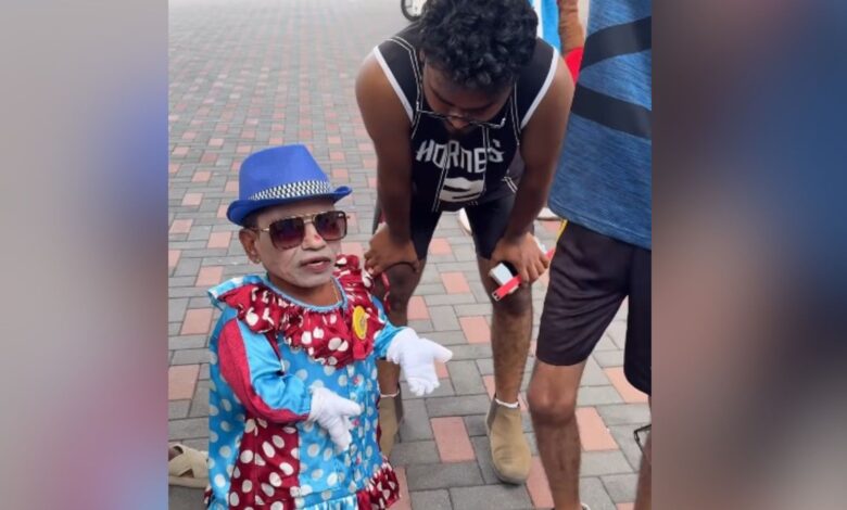 Child dressed as a clown with sunglasses and a blue hat, standing outdoors on a brick pavement.