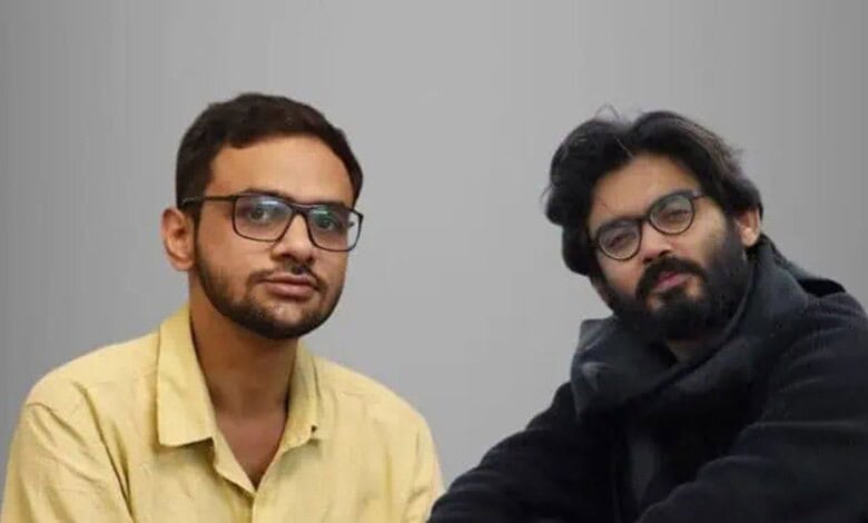 Two men with glasses sitting against a plain grey background.