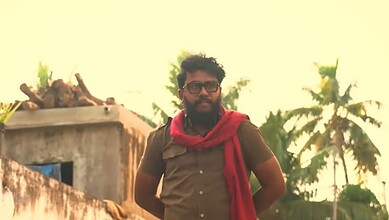 Man with glasses and beard wearing a red scarf in front of palm trees at sunset.