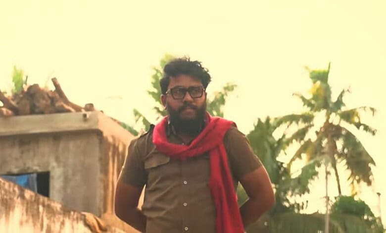 Man with glasses and beard wearing a red scarf in front of palm trees at sunset.