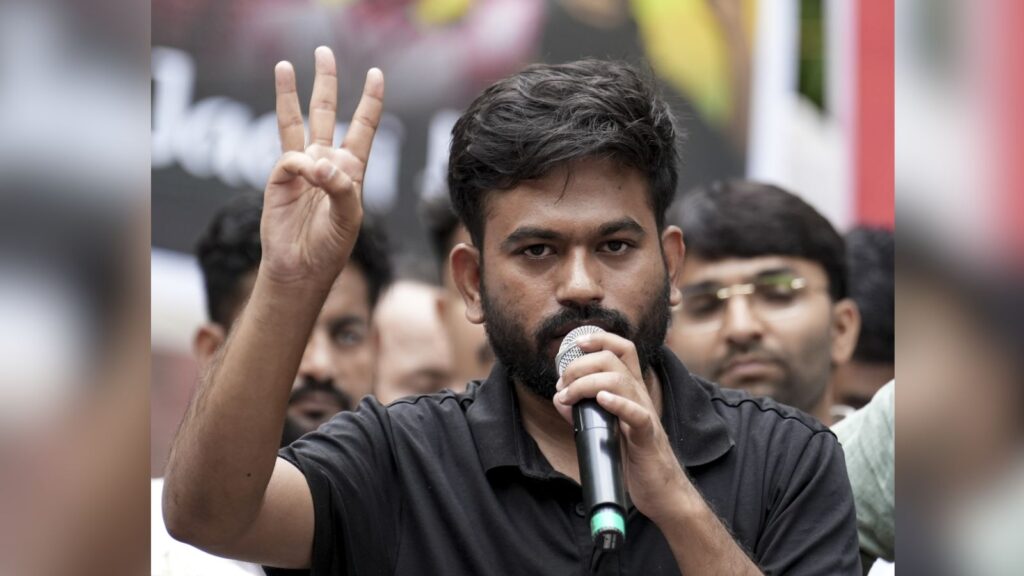 Protestor with beard holding microphone and making a V sign at a rally.