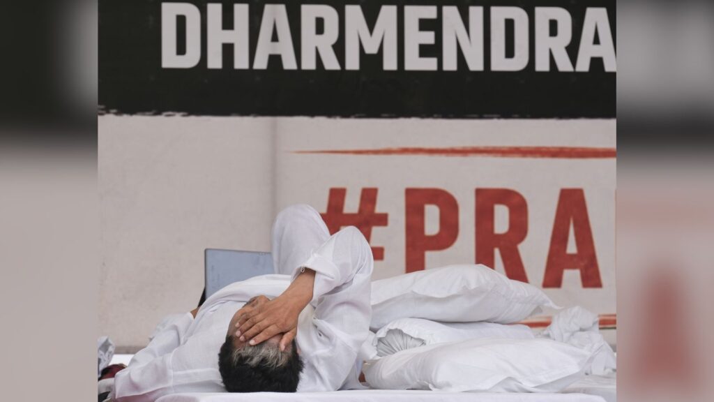 Person resting with head in hand on a bed with white sheets, protest banners in background.