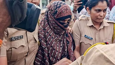 Nida Khan in police custody with officers around her, wearing a headscarf and glasses.
