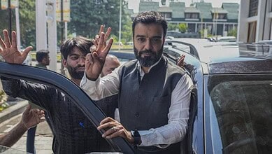 Man with beard and white shirt waving to the camera outside a vehicle.
