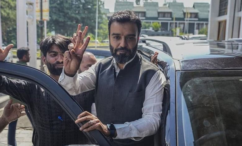 Man with beard and white shirt waving to the camera outside a vehicle.