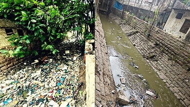 Polluted water canal with trash and debris in an urban area, surrounded by brick walls and greenery.