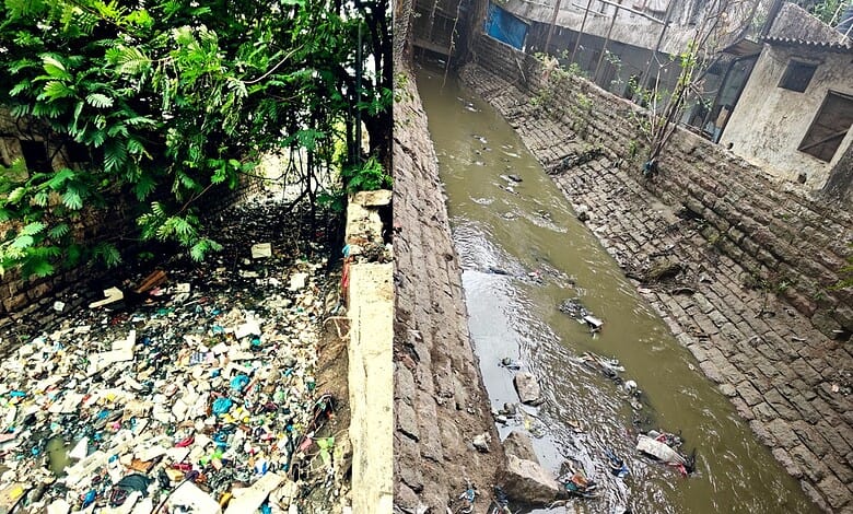 Polluted water canal with trash and debris in an urban area, surrounded by brick walls and greenery.