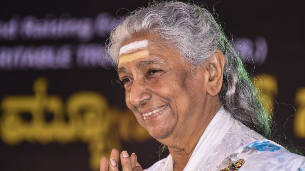 Elderly woman with grey hair smiling, wearing traditional attire and a white mark on her forehead.
