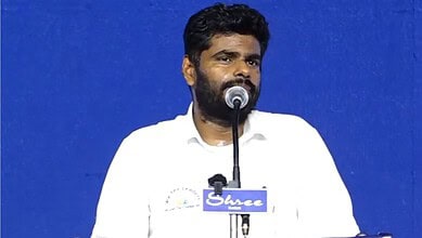 Annamalai speaking at a public event with a blue background and microphone.