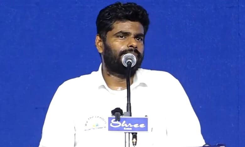 Annamalai speaking at a public event with a blue background and microphone.