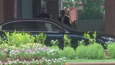 Two men in suits near a black car surrounded by green plants and flowers.