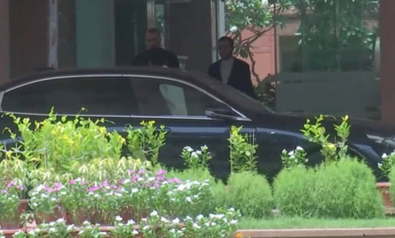 Two men in suits near a black car surrounded by green plants and flowers.