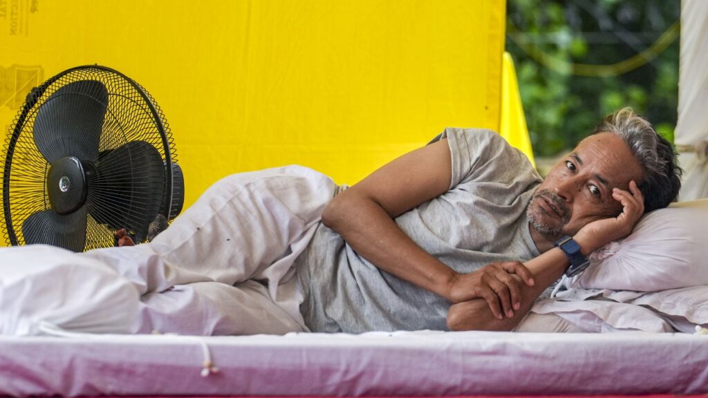 Man lying on bed in a yellow tent, appearing in pain and resting his head on his hand.