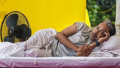Man lying on bed in a yellow tent, appearing in pain and resting his head on his hand.