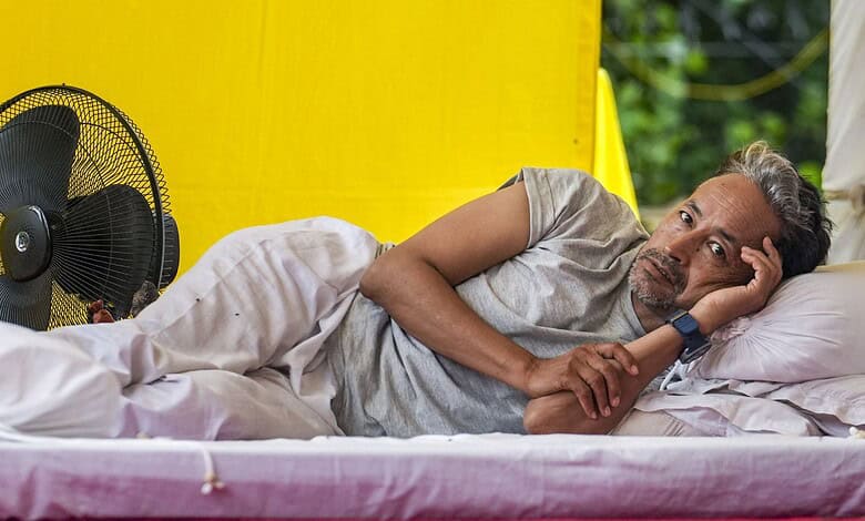 Man lying on bed in a yellow tent, appearing in pain and resting his head on his hand.