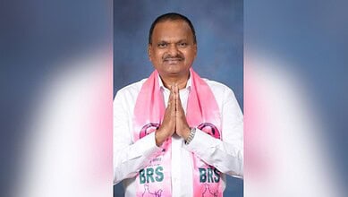 Smiling man in white kurta with pink scarf, hands folded in greeting, against blue background.
