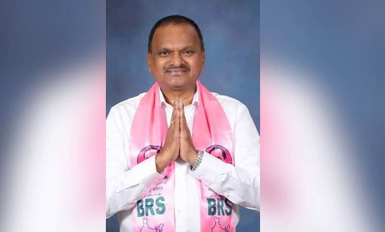 Smiling man in white kurta with pink scarf, hands folded in greeting, against blue background.