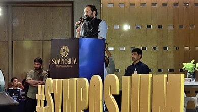 Uttam at the symposium speaking at a podium with the hashtag #SYMPOSIUM and a large golden sign.