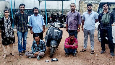 Group of nine men and two boys standing outdoors with a scooter and motorcycles in the background.