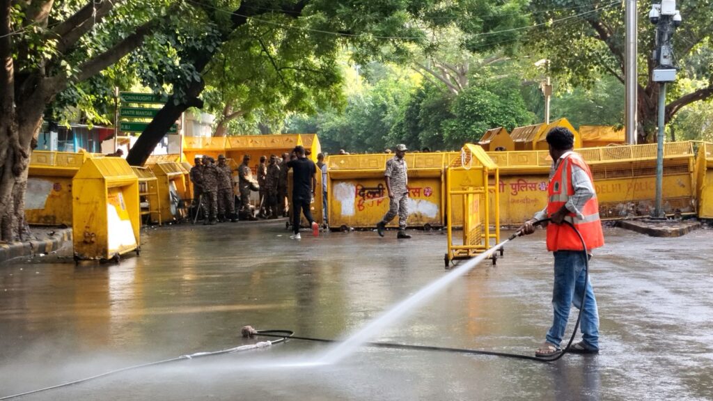 Man cleaning a wet street near barricades and police officers at Jantar Mantar, India.