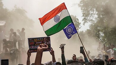 Person holding Indian flag at rally with crowd and smoke in background.