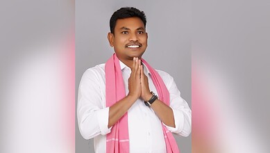 Smiling man in white shirt with pink scarf, hands folded in greeting, on grey background.