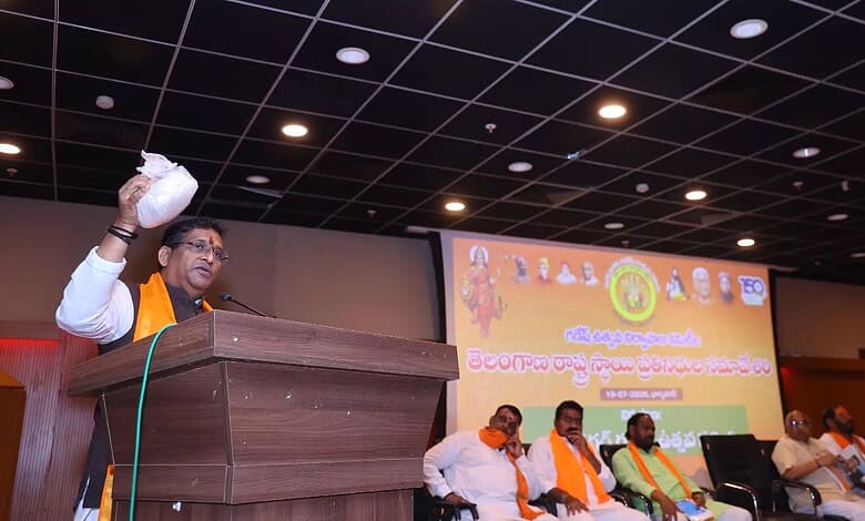 Speaker at Khairatabad Ganesh event holding a white bag, with a large screen behind him.