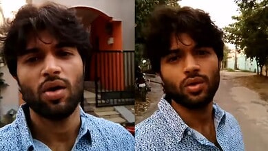 Young man with dark hair and beard wearing a blue patterned shirt outdoors.