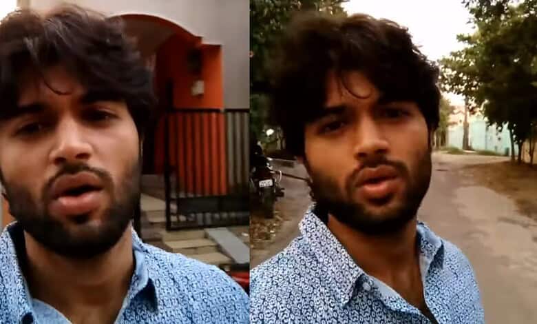 Young man with dark hair and beard wearing a blue patterned shirt outdoors.
