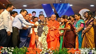Group of people, including monks and officials, at a ceremonial event with flowers and a large screen in.