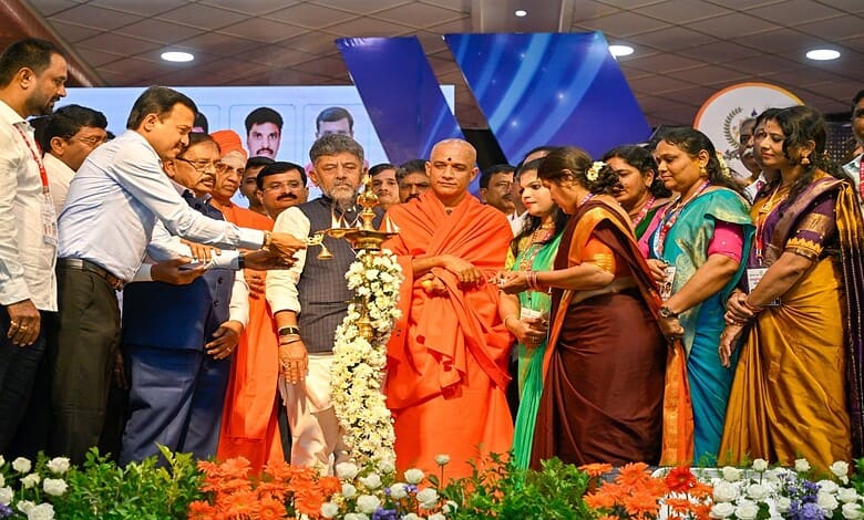 Group of people, including monks and officials, at a ceremonial event with flowers and a large screen in.