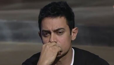 A man with short black hair and a serious expression, resting his chin on his hand.