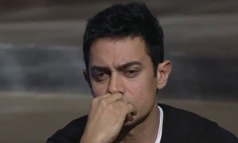 A man with short black hair and a serious expression, resting his chin on his hand.