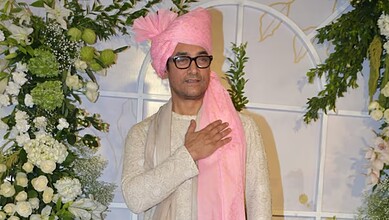 Indian man in traditional attire with pink turban and glasses at a wedding event.