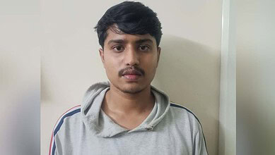 Young man with serious expression, wearing grey hoodie, standing against plain wall.