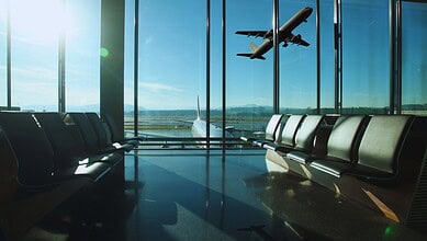 Empty airport departure lounge with rows of seating overlooking a runway as a plane takes off into a clear blue sky