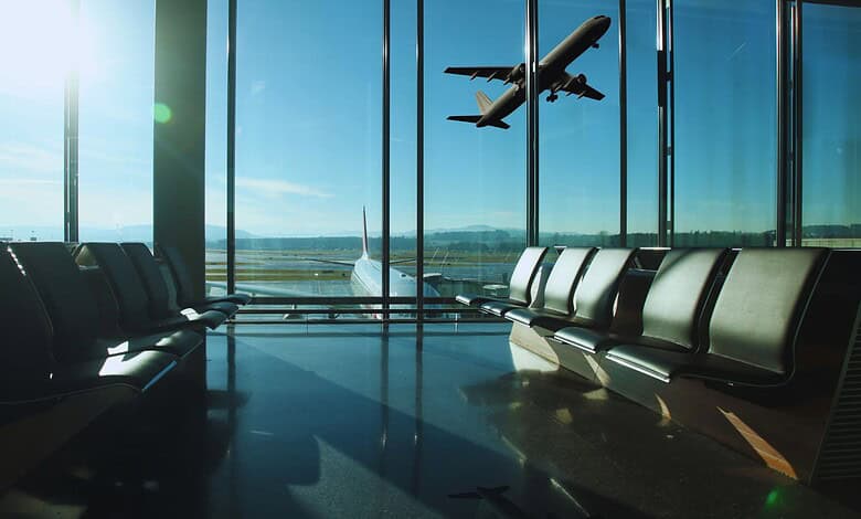 Empty airport departure lounge with rows of seating overlooking a runway as a plane takes off into a clear blue sky