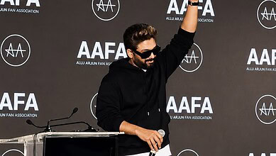 Allu Arjun at Hyderabad event, wearing sunglasses and black hoodie, speaking on stage.