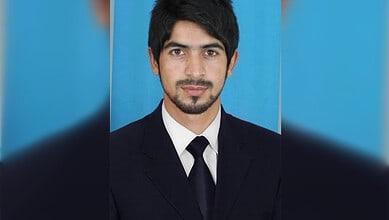 Young man in formal suit with a blue background, looking confidently at the camera.