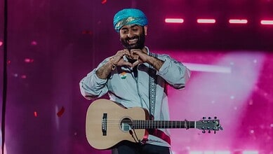 Arijit Singh performing on stage with a guitar and a blue turban, smiling and making a heart shape with.