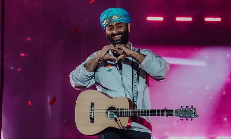 Arijit Singh performing on stage with a guitar and a blue turban, smiling and making a heart shape with.