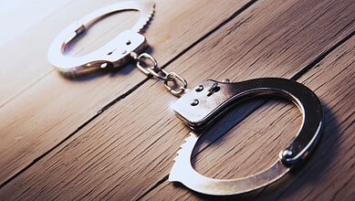 A pair of open metal handcuffs lying on a wooden surface, connected by a chain.