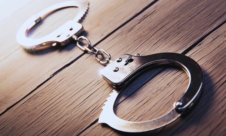 A pair of open metal handcuffs lying on a wooden surface, connected by a chain.