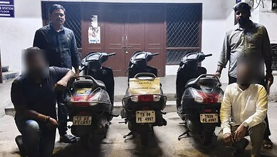 Three scooters with similar number plates parked in front of a building with four men standing nearby.
