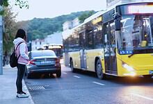 Woman waiting at a bus stop as a yellow city bus approaches on a tree-lined street at dusk.