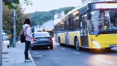 Woman waiting at a bus stop as a yellow city bus approaches on a tree-lined street at dusk.