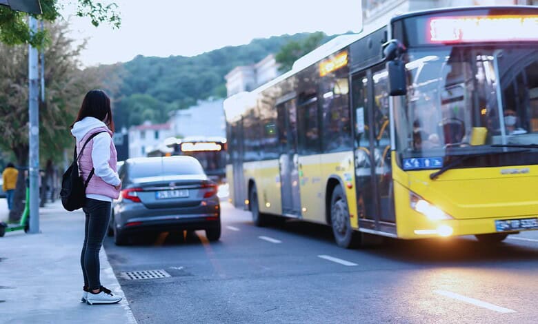 Woman waiting at a bus stop as a yellow city bus approaches on a tree-lined street at dusk.