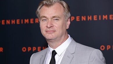 Christopher Nolan in a grey suit at a film event, with 'Oppenheimer' backdrop.